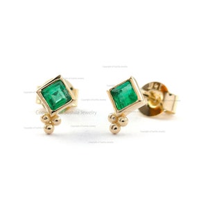 May include: A pair of gold earrings with small emerald green square gemstones and three small gold balls on each earring.