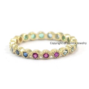 May include: A gold band ring featuring a row of colourful gemstones. The gemstones are set in a scalloped design and include blue, green, and pink stones.