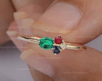 Op de afbeelding: Een delicate gouden ring met een ovale smaragd, een ronde robijn, een ronde saffier en drie kleine diamanten. De edelstenen zijn gegroepeerd op de ringband, waardoor een uniek en elegant ontwerp ontstaat. De ring wordt door een persoon vastgehouden.