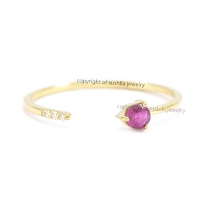 May include: A delicate gold ring with a split shank design. The ring features a small, round, bright pink gemstone on one end and three small white gemstones on the other. The ring is a minimalist style.
