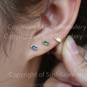 May include: Three gold bar earrings with different colored gemstones. The earrings are shown on a person's earlobe. The gemstones are blue, green, and clear.
