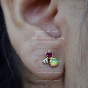 May include: A close-up of a gold earring featuring three gemstones: a red ruby, a clear diamond, and a green opal. The earring is set in a simple, modern style.