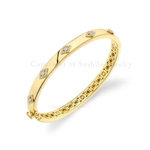 Puede incluir: Un brazalete de oro con un diseño delicado e intrincado. El brazalete está adornado con pequeños diamantes transparentes engastados en un patrón geométrico. El brazalete está abierto y tiene un cierre con bisagra.