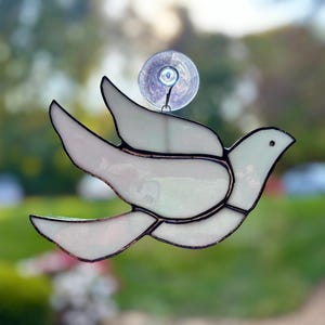 May include: A white stained glass dove with a suction cup for hanging on a window. The dove is in flight with its wings spread.