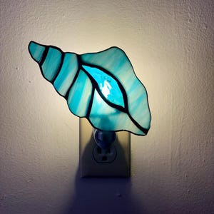 May include: A stained glass night light shaped like a seashell. The shell is a mix of light and dark blue with black outlines. The night light is plugged into a wall outlet and emits a soft glow.