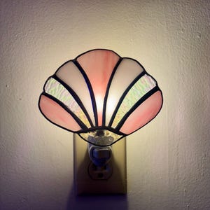 May include: A seashell-shaped night light constructed from stained glass panels in shades of soft pink and iridescent white. The glass pieces are joined by dark metal soldering, creating a fan-like pattern that glows when plugged into a wall outlet.