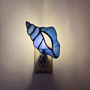 May include: A stained glass night light shaped like a seashell, featuring various shades of blue and white glass pieces joined by dark metal solder lines. The light emits a soft glow from behind the shell, which is plugged into a wall outlet.