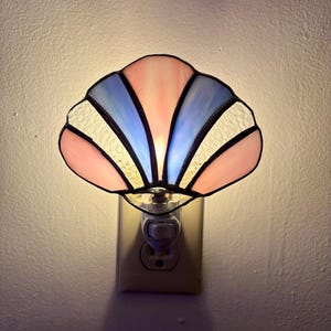 May include: A stained glass night light shaped like a seashell with alternating panels of pale pink, light blue, and textured white glass. The lamp is plugged into a wall outlet and emits a soft, warm glow through the translucent glass segments.
