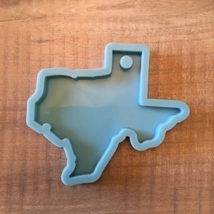Puede incluir: Un molde de silicona azul claro con forma del estado de Texas. El molde tiene un pequeño agujero en la parte superior para colgarlo.