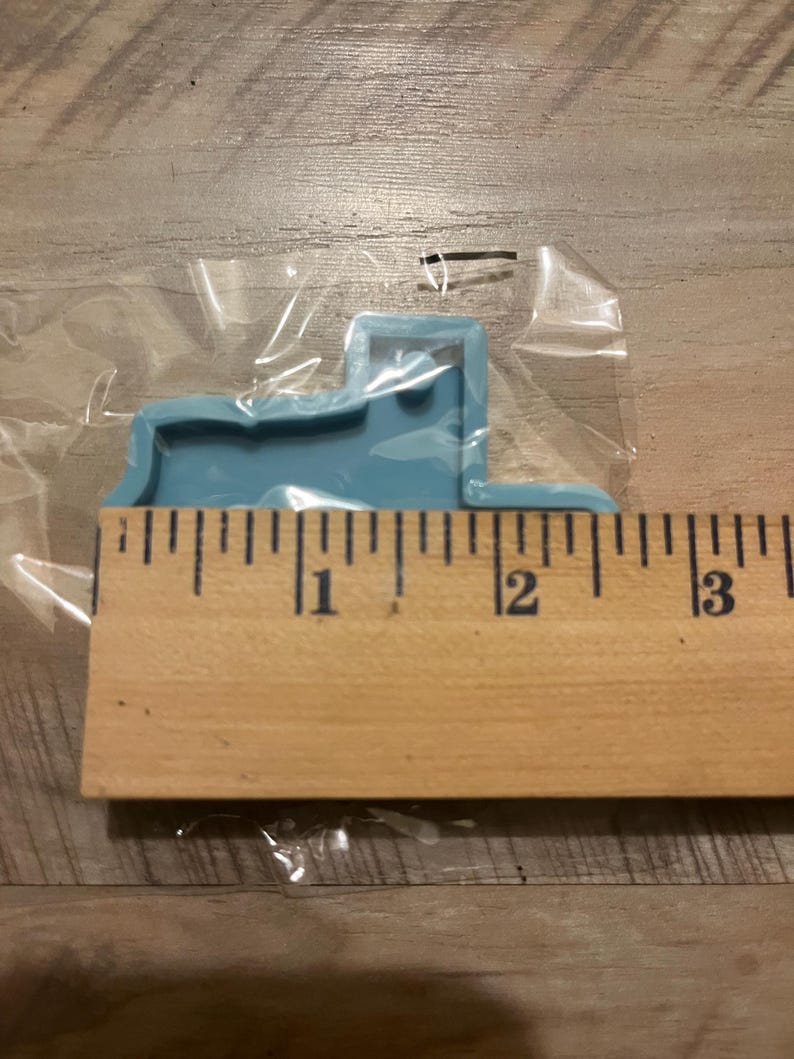 May include: A light blue silicone mould shaped like a truck. The mould is approximately 2.5 cm wide and 5 cm long. The mould is sitting on a wooden ruler.