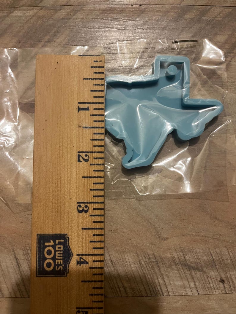 May include: A light blue plastic Texas state cookie cutter measuring approximately 10 cm in length. The cutter is on a wooden surface with a ruler for scale.