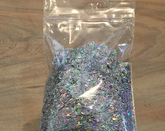 Puede incluir: Una bolsa de plástico transparente llena de purpurina plateada iridiscente. La purpurina tiene forma de pequeños rectángulos delgados.