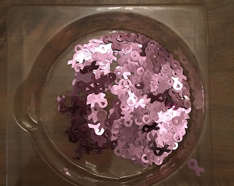 May include: A clear plastic container filled with pink glitter shaped like ribbons. The container is on a wooden surface.