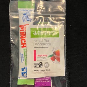 May include: A clear plastic bag containing three packets of dietary supplements. One packet is green and white with the text "PUNCH TROPICAL" and "To Go!". Another packet is white with "Herbal Tea Concentrate" and "raspberry" text. The third packet is purple.