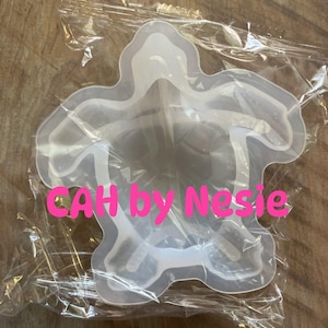 Puede incluir: Un molde de silicona translúcido y flexible con forma de tortuga marina, envuelto en una bolsa de plástico transparente. El texto CAH by Nesie está impreso en letras de color rosa brillante en el centro del molde, que tiene una cavidad interior.
