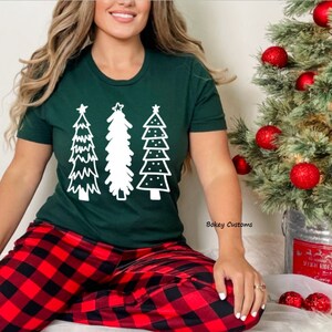 May include: A green t-shirt with a white design of three stylized Christmas trees. The person wearing the shirt is sitting on a red and black plaid blanket.