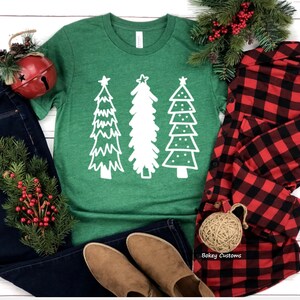 May include: A green t-shirt with a white design of three stylized Christmas trees. The trees have a star on top and are decorated with dots and lines. The t-shirt is laid flat on a white wooden surface with a pair of brown boots, blue jeans, a red and black plaid shirt, and a small red bell ornament.