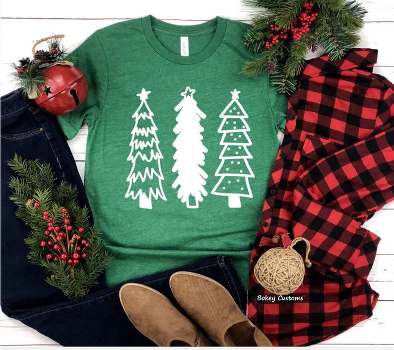 May include: A green t-shirt with a white design of three stylized Christmas trees. The trees have a star on top and are decorated with dots and lines. The t-shirt is laid flat on a white wooden surface with a pair of brown boots, blue jeans, a red and black plaid shirt, and a small red bell ornament.