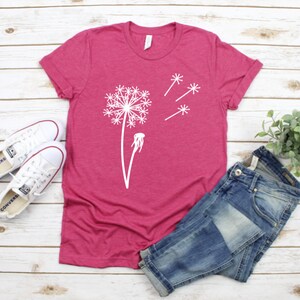 以下が含まれることがあります： 白いタンポポのグラフィックが描かれたピンクのTシャツ。タンポポは種を風に乗せて飛ばしています。Tシャツは白い木製の表面に置かれ、白いスニーカーと青いジーンズが置かれています。