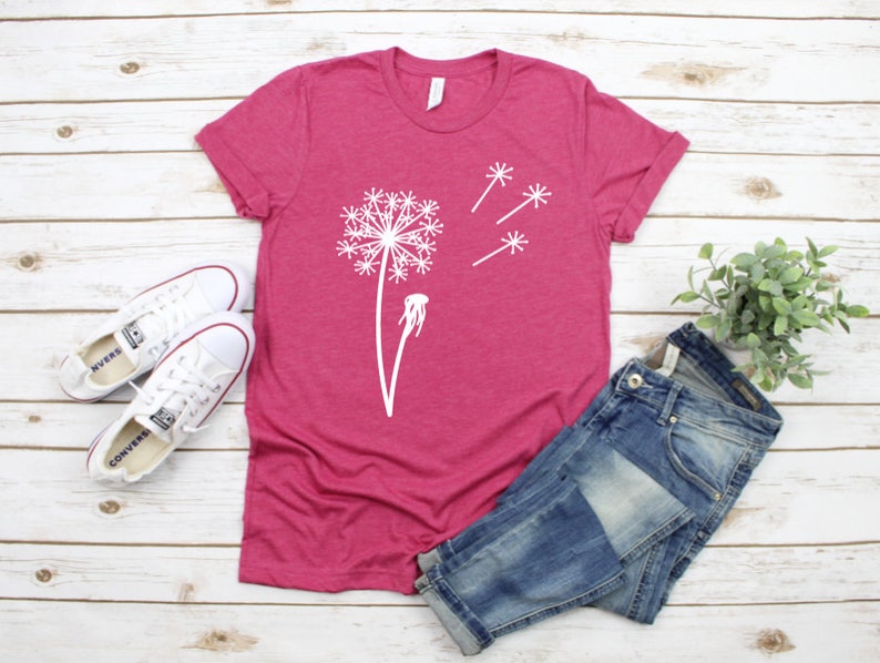 以下が含まれることがあります： 白いタンポポのグラフィックが描かれたピンクのTシャツ。タンポポは種を風に乗せて飛ばしています。Tシャツは白い木製の表面に置かれ、白いスニーカーと青いジーンズが置かれています。