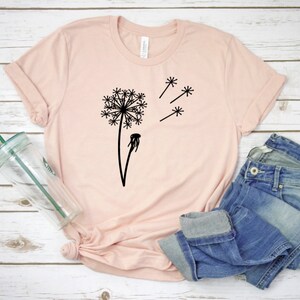 以下が含まれることがあります： 白い木製の表面に置かれた薄いピンクのTシャツ。黒いタンポポのグラフィックが描かれており、タンポポの種が風に乗って飛んでいる。透明なガラスのタンブラーと青いジーンズも置かれている