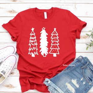 May include: Red t-shirt with a white design of three stylized Christmas trees. The shirt is laying on a white wooden surface with a pair of white sneakers and blue denim shorts.