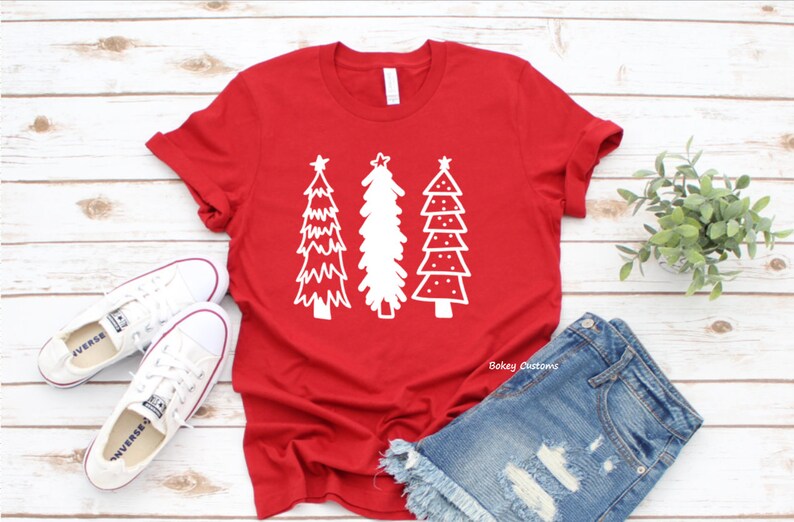 May include: Red t-shirt with a white design of three stylized Christmas trees. The shirt is laying on a white wooden surface with a pair of white sneakers and blue denim shorts.