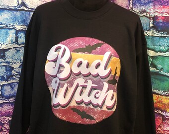 May include: Black sweatshirt with a circular graphic that reads "Bad Witch" in a retro font. The graphic features a sunset design with bats. The sweatshirt is displayed on a black mannequin.