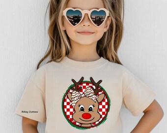 May include: A beige t-shirt featuring a cartoon reindeer design with a red nose, antlers, and a bow. The reindeer is set within a red and white checkered circle, framed by a green border. The text "Bakey Customs" is printed on the shirt.