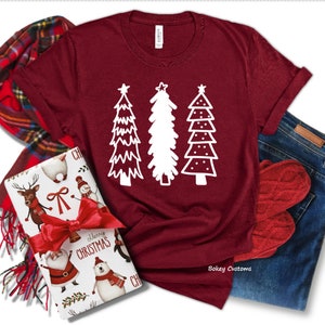 May include: A maroon t-shirt with a white design of three stylized Christmas trees. The trees are decorated with stars and dots. The shirt is laid flat on a white background.