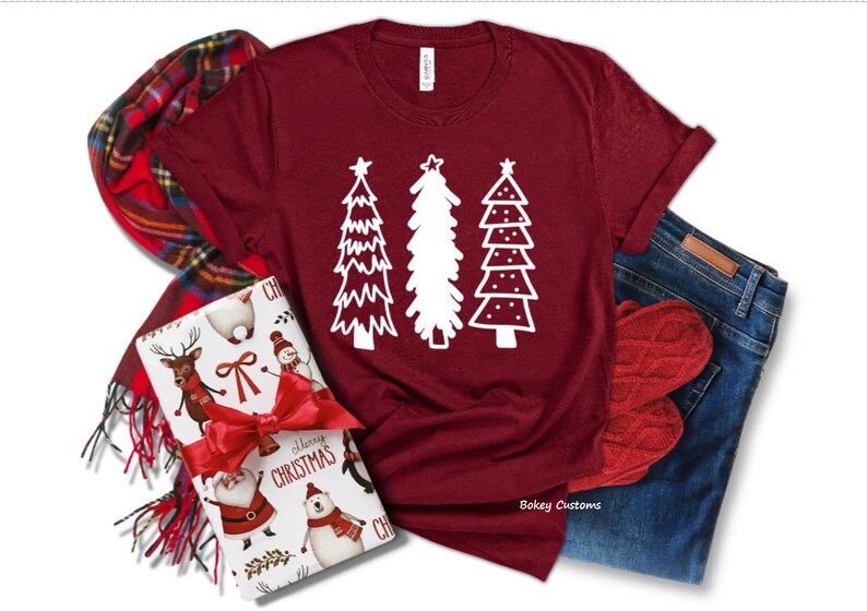May include: A maroon t-shirt with a white design of three stylized Christmas trees. The trees are decorated with stars and dots. The shirt is laid flat on a white background.