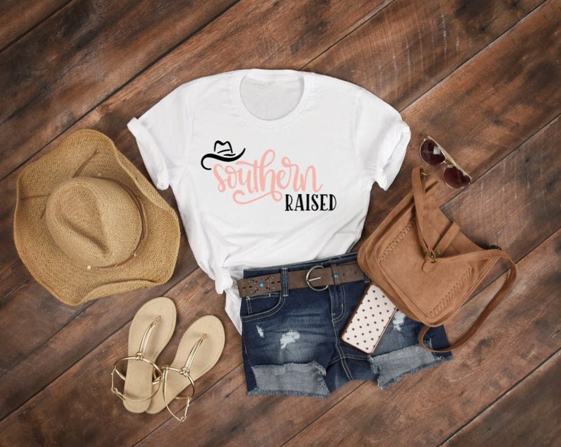 May include: White t-shirt with a pink and black graphic that says "Southern Raised" with a cowboy hat design. The shirt is laid out on a wooden surface with a straw hat, sandals, a denim skirt, and a brown leather purse.