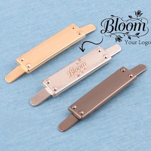 May include: Three rectangular metal bag tags with prongs, featuring a polished gold finish, a brushed silver finish with the engraved text Bloom, and a matte bronze finish. Each tag has two small rivets on the front surface for attachment.