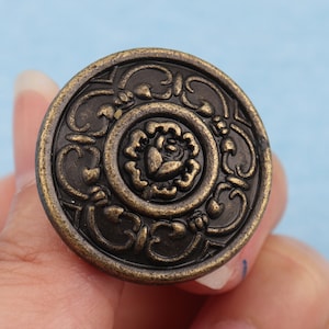 May include: A round, antique brass-colored cabinet knob with a floral design. The knob features a raised rose in the center, surrounded by ornate scrollwork. The edges have a slightly darker patina, giving it a vintage look.