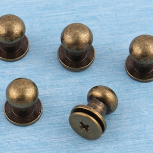 May include: Five antique gold-toned metal knobs with a round top and a flat base. The knobs have a screw-on design.