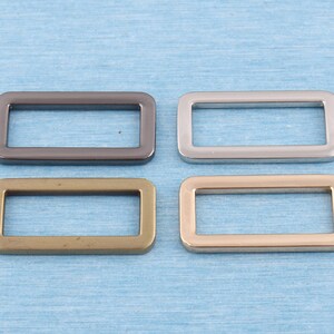 May include: Four metal D-rings in different finishes: black, silver, gold, and antique brass. The rings are rectangular with rounded corners and a smooth surface.