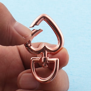 Könnte beinhalten: Roségoldener Herz-förmiger Auslöseverschluss aus Metall. Der Verschluss hat einen federbelasteten Verschluss und einen drehbaren Haken.