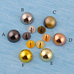 May include: A collection of decorative metal rivets in various colors, including black, silver, gold, and copper. The rivets have a rounded, dome-like shape and are designed for attaching to fabric or leather. The image shows the rivets with their corresponding posts.