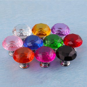 Op de afbeelding: Een verzameling van tien facetgeslepen kristallen kastknoppen in verschillende kleuren, waaronder helder, geel, paars, rood, groen, roze, blauw, oranje en zwart. Elke knop heeft een zilverkleurige basis.