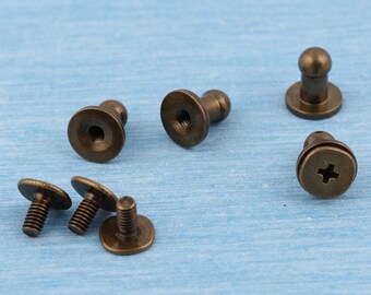 May include: A set of six antique gold-toned metal fasteners. The fasteners have a round head with a raised center and a screw-on base. Some of the fasteners have a small knob on top of the head.