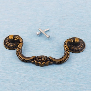 May include: Antique brass drawer pull with decorative detailing. The pull has a curved handle and two round mounting plates. Two silver screws are included. The hardware is designed for furniture restoration or new builds.