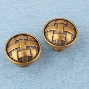 Peut inclure: Deux boutons de meuble en laiton antique avec un motif tressé sur une surface bleu clair. Les boutons sont ronds, avec un dessus légèrement bombé et une courte tige. Le laiton a une finition vieillie et chaleureuse.