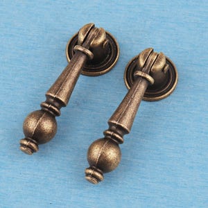 May include: Two antique brass-finished metal drop pulls featuring a circular mounting plate and a vertical, tapered handle with a spherical bottom finial. These hardware pieces have a textured, weathered appearance with dark brown highlights.