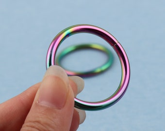 May include: Two iridescent metal rings, one held in a hand, the other lying on a blue surface. The rings are a mix of pink, green, and blue.