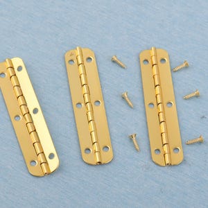Op de afbeelding: Drie goudkleurige scharnieren met afgeronde uiteinden en meerdere schroefgaten liggen op een lichtblauwe ondergrond. Verschillende kleine gouden schroeven liggen verspreid rond de scharnieren.