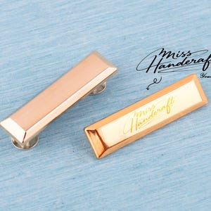 May include: Two rectangular cabinet pulls in a rose gold finish with silver-toned bases. One pull has the text "Miss Handcraft" in a cursive font. The other pull is blank. The background is a light blue.
