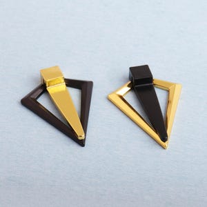 May include: Two geometric earrings. One earring features a gold-colored triangle frame with a brown center and a gold-colored rectangular top. The other earring has a gold-colored triangle frame with a black center and a black rectangular top.