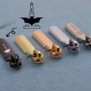 Könnte beinhalten: Eine Auswahl an Metall-Reißverschluss-Anhängern in Gold, Silber und Bronze. Jeder Anhänger hat eine rechteckige Oberseite mit einem dekorativen Design und eine Schlaufe zum Befestigen an einem Reißverschluss. Das Bild enthält auch ein Logo des Eiffelturms und einer Fledermaus.