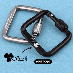 Könnte beinhalten: Zwei quadratische Karabiner, einer in Silber und einer in Schwarz. Der silberne Karabiner ist mit dem Wort STRONG graviert. Der schwarze Karabiner hat einen Schraubverschluss, ein Kleeblatt-Symbol und das Wort Luck. Unten stehen ein Kleeblatt und Luck.