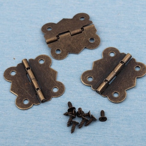 Può includere: Tre cerniere color bronzo antico con bordi decorativi e fori per viti sono esposte su una superficie azzurra. Sono presenti anche diverse piccole viti in bronzo. Queste cerniere sono adatte a piccole scatole o lavori artigianali.